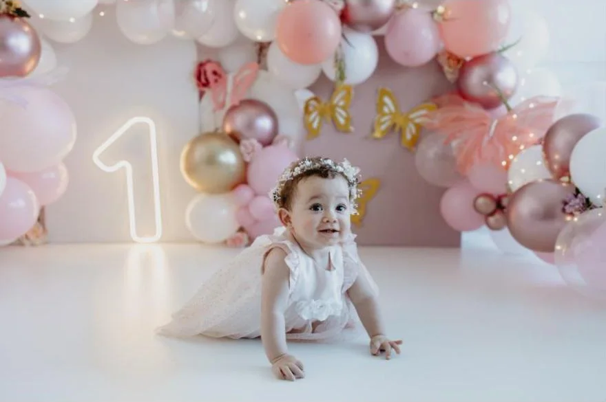 Bebê de vestido branco e coroa de flores engatinhando em cenário de balões rosa